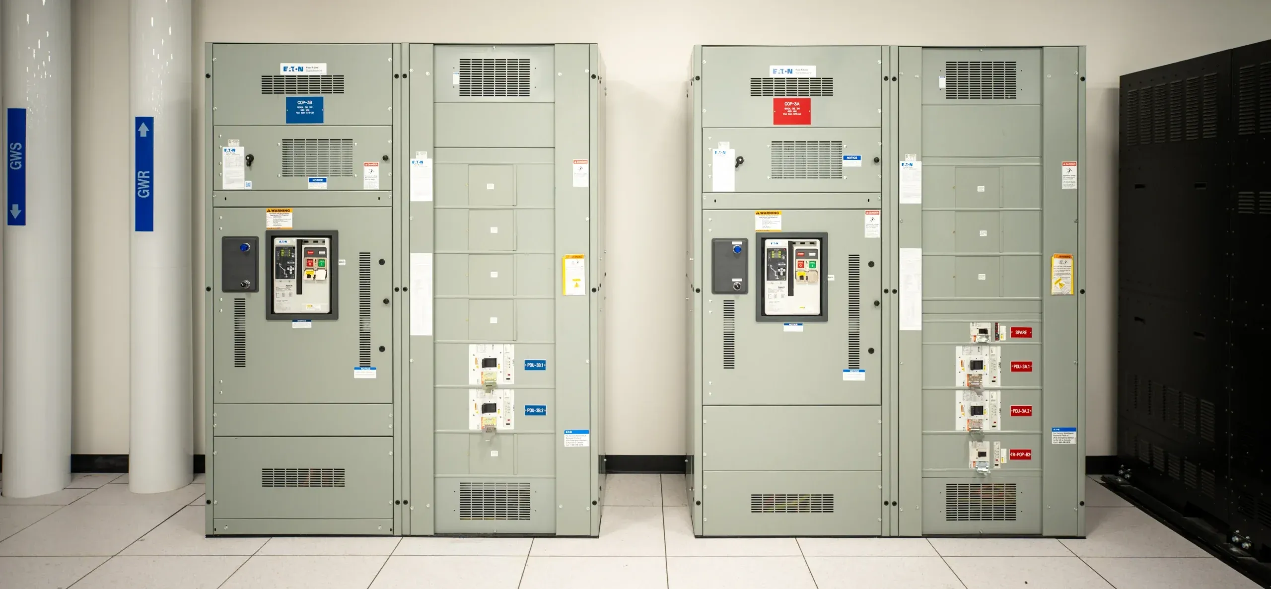 Rows of gray electrical gear cabinets in a data room, labeled breakers and controls, with blue safety signs on white columns left side.