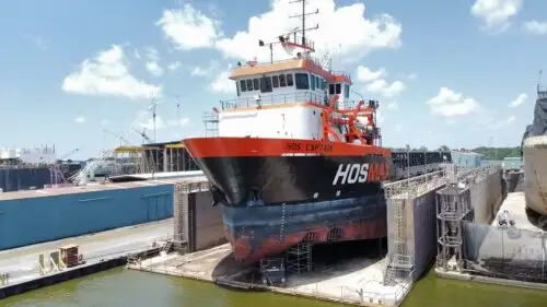 Tugboat in a dry dock undergoing maintenance at a harbor, raised above the water with blue gates and ladders nearby.